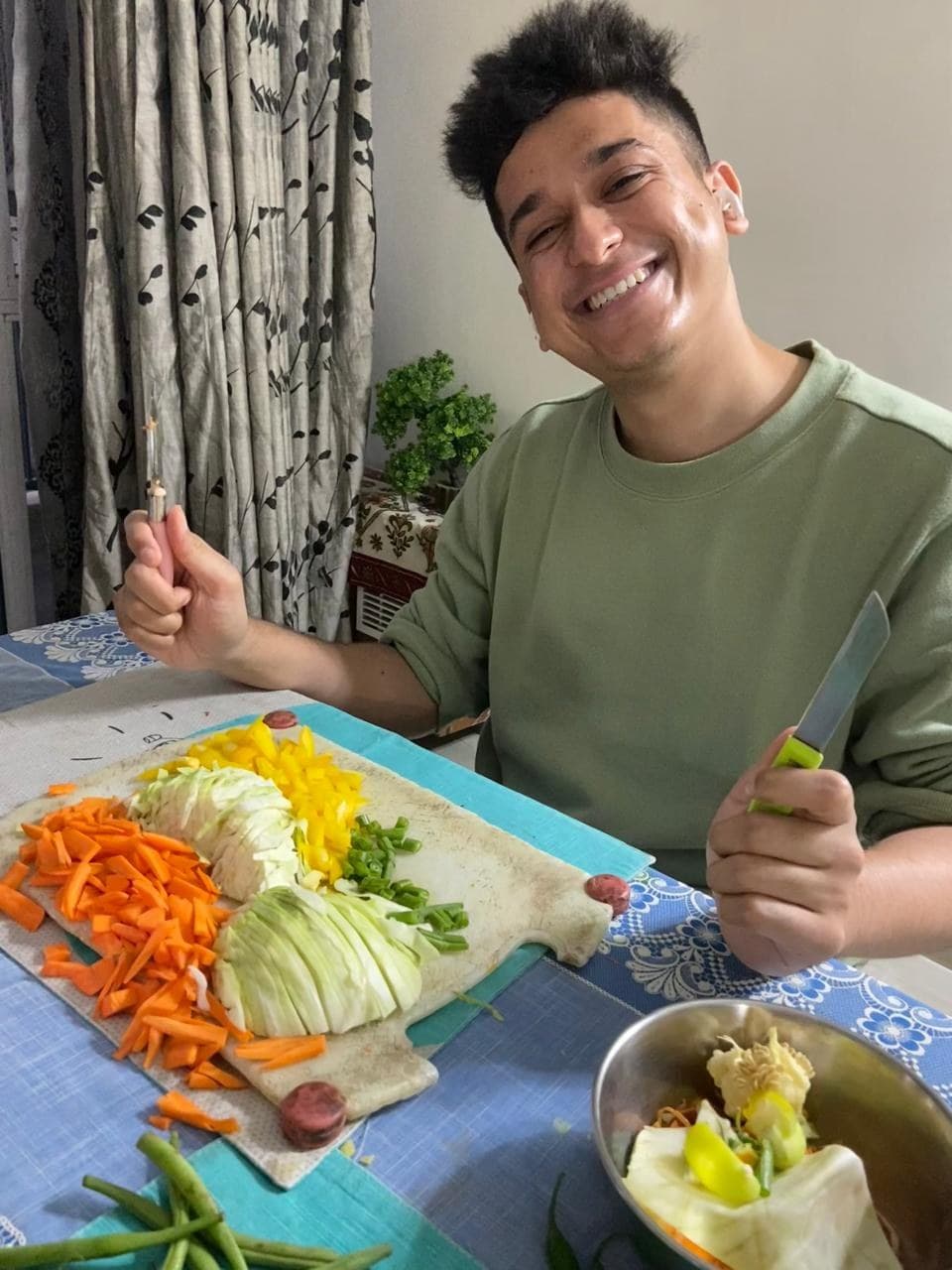 Kushagra at a table mid-preparation, knife in hand, laughing, with julienned carrot, cabbage, peppers and beans laid out in neat rows on a board.
