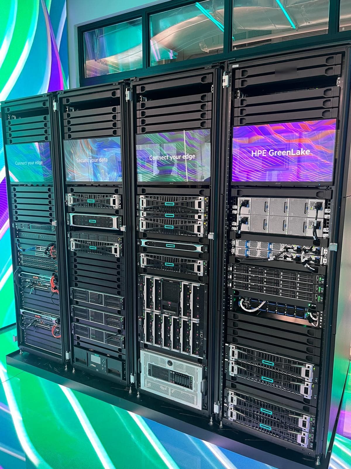Four racks of HPE servers, storage and network switches, with display screens mounted between the equipment.