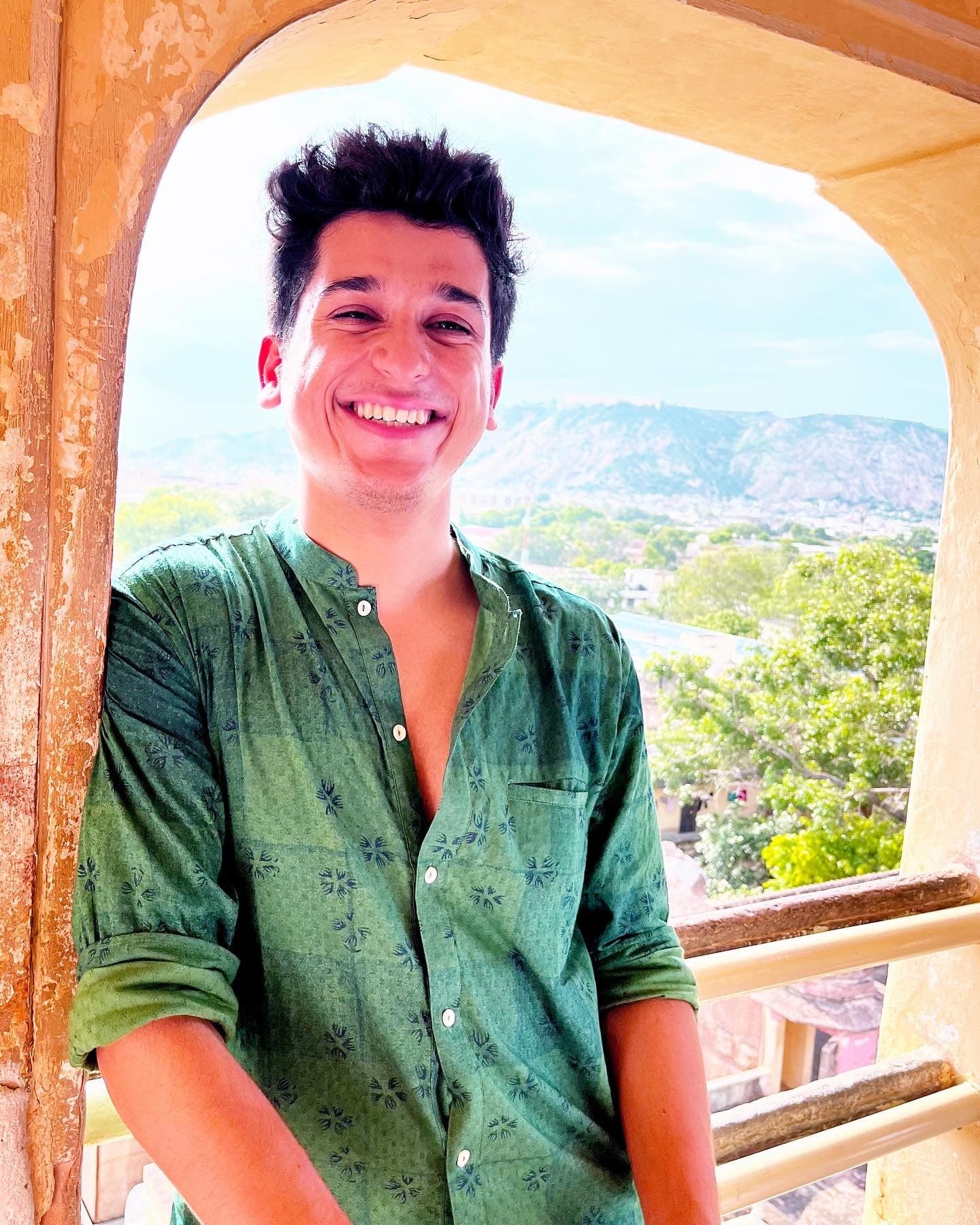 Kushagra framed by a sandstone arch, a hill town behind him, in a green kurta, in natural light.