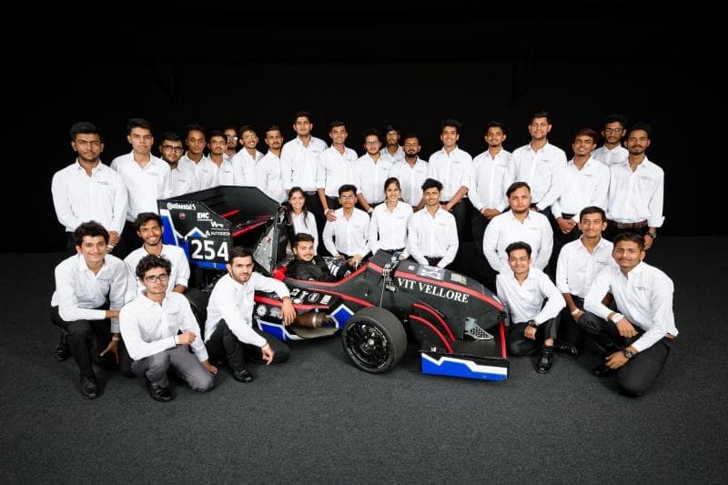 The full Pravega Racing team of about thirty-five people in white shirts, arranged around their Formula Student car under studio lighting on black.